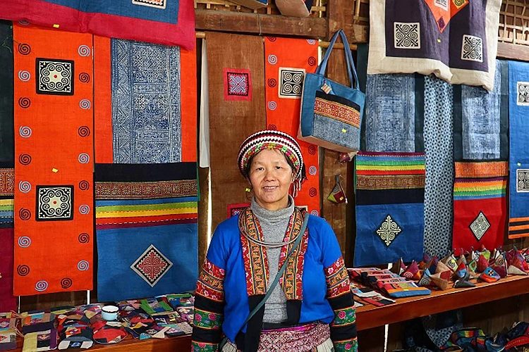 An artisan in traditional Hmong dress at the Lung Tam linen weaving village, Ha Giang, surrounded by indigo and embroidered textiles