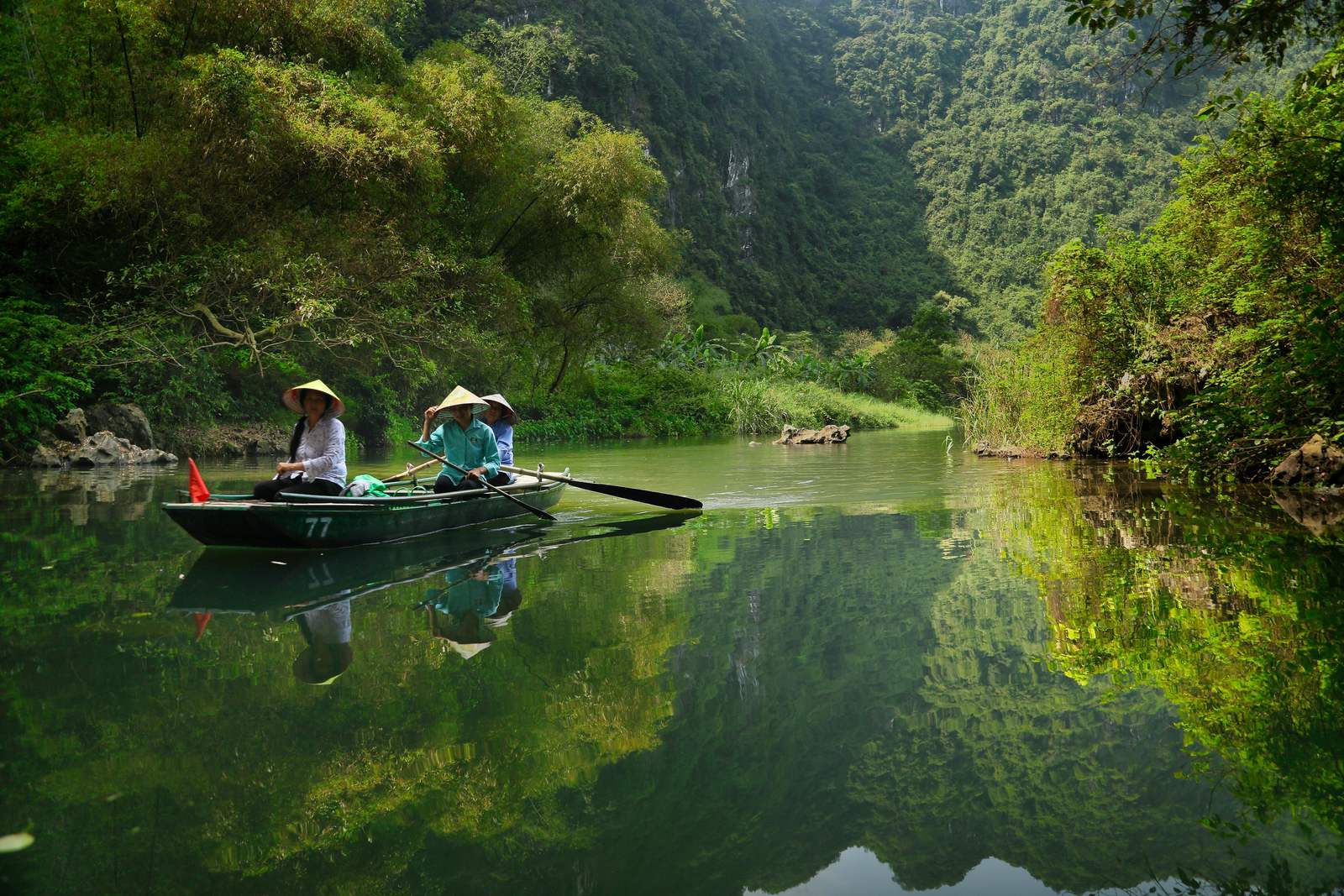 Two travellers in a rowing boat on the river at Trang An, Ninh Binh, between green limestone cliffs